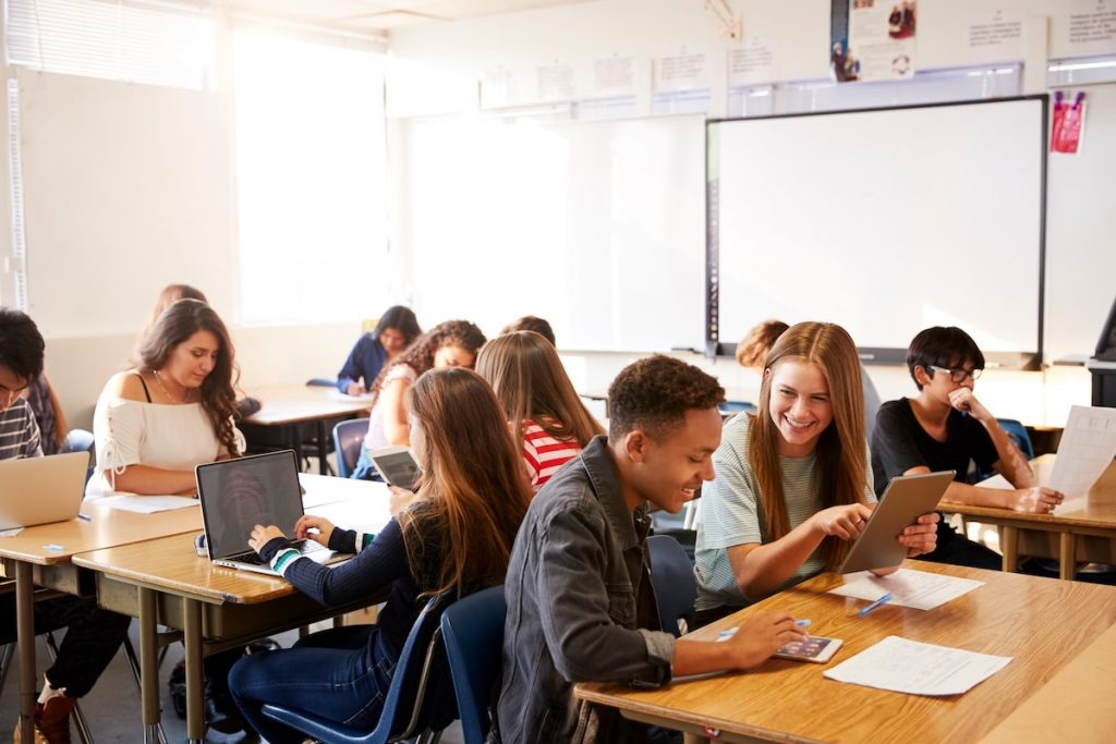 high school students sit in a classroom during class time, looking at laptops, tablets, and paperwork at their desks.